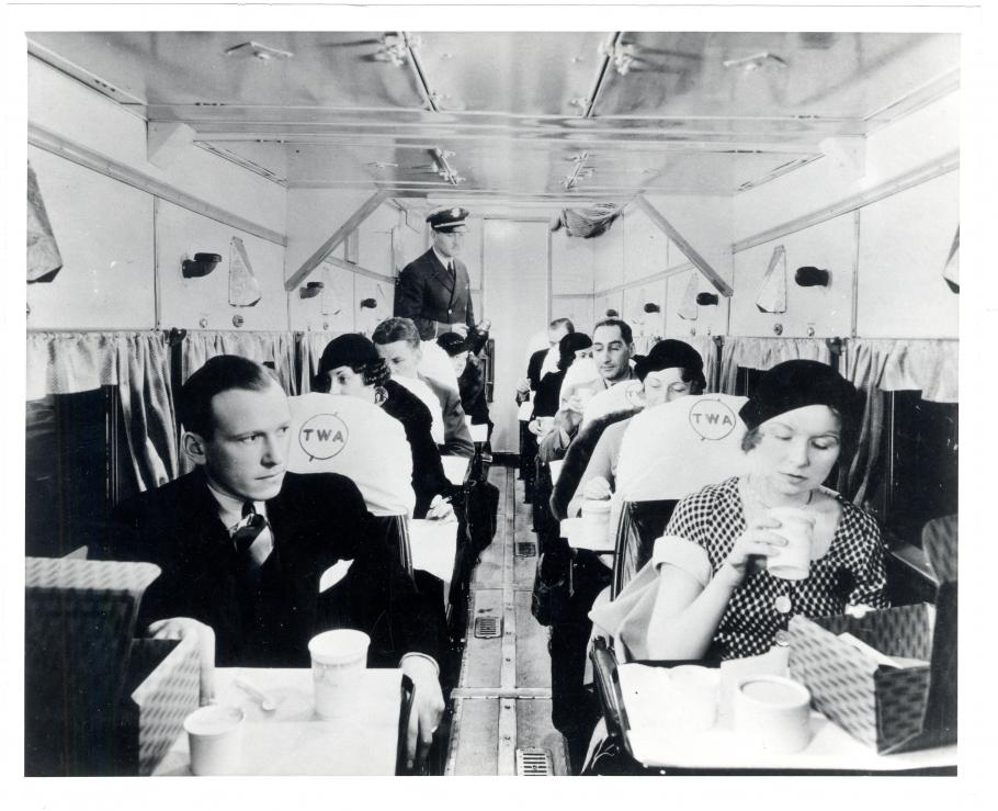 Interior of an early commercial aircraft. People sit in seats that have two rows of one seat apiece. The people in the front seem to have received some form of food service.