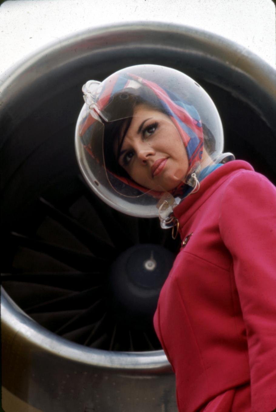 Side view of a flight attendant wearing a red flight uniform and a plastic, bubble-shaped helmet.