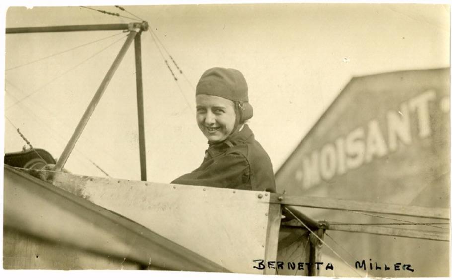 Woman in flying headgear sits in the cockpit of an early airplane. Text in bottom right reads: "Bernetta Miller."