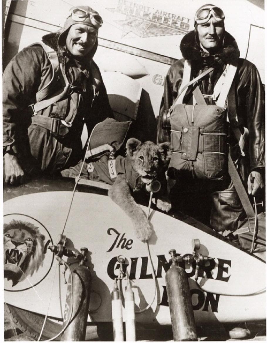 Two men in flight gear pose behind a lion cub at center, who also appears to be wearing flight gear.