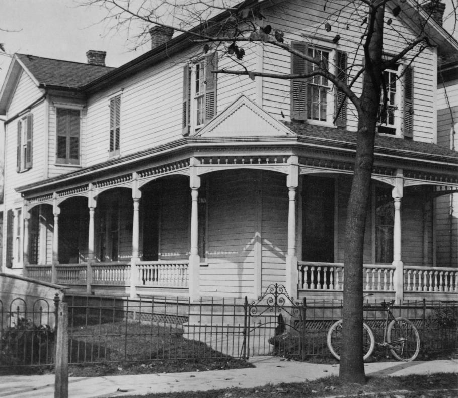 A large white house with a wrap around porch. A bicycle is leaned up against a short iron fence which surrounds the yard.