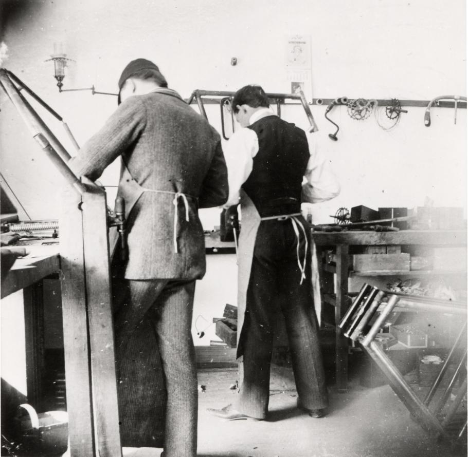 Two men with their back turned to the camera, working at a work bench and surrounded by tools.