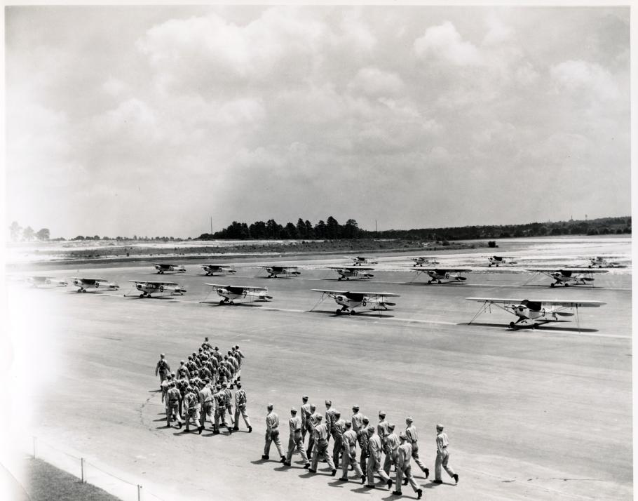 Multiple lines of Piper Cubs, monoplanes with one engine, are seen on a U.S. Navy training site. Groups of men are walking in formation in front of the planes.