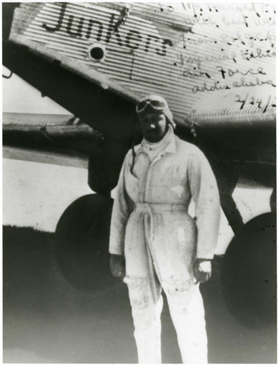 Black man in flight suit and leather pilot's helmet stands in front of left side of nose of plane made of corrugated metal, Junkers in capital letters over his head. Handwritten note in upper right.