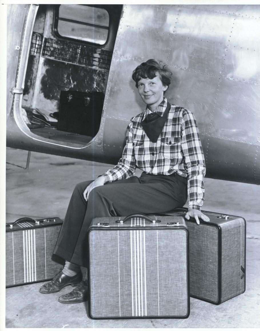 Amelia Earhart, a white female pilot, poses in front of an aircraft with three pieces of luggage that she designed.