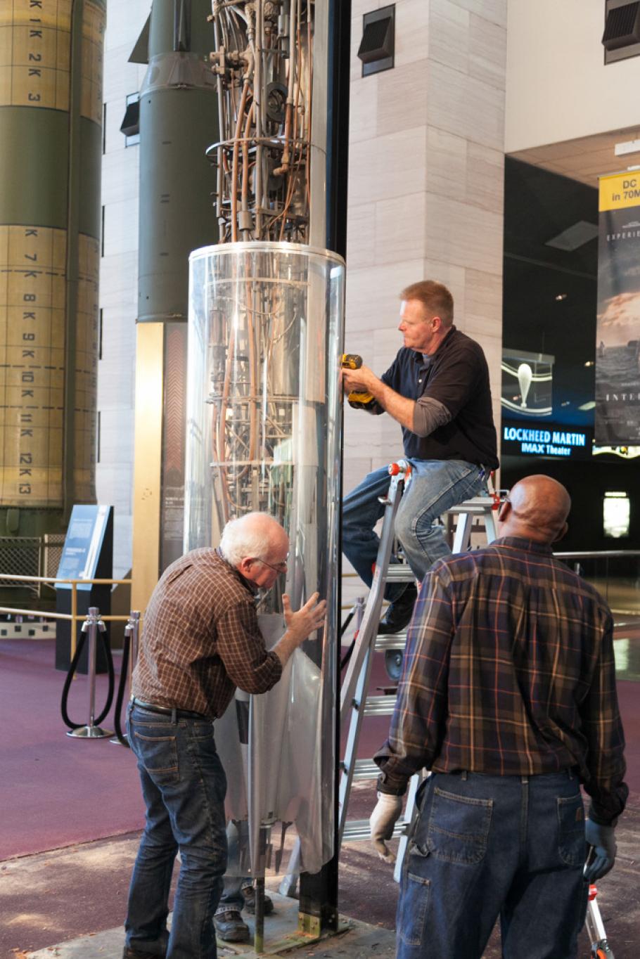 Three Museum staff members work to remove a 1940s rocket from a protective case prior to transporting the rocket.