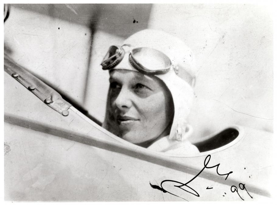 Amelia Earhart sits inside the cockpit of an aircraft. She is wearing an aviator hat and goggles.
