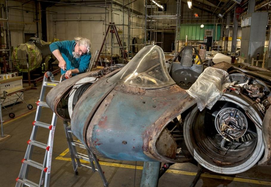 A white, male aircraft mechanic performs work on a gray and white metal aircraft undergoing conservation treatment.