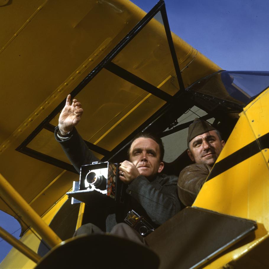 A photo taken from underneath an aircraft. One man leans out with a camera, the other is a pilot.