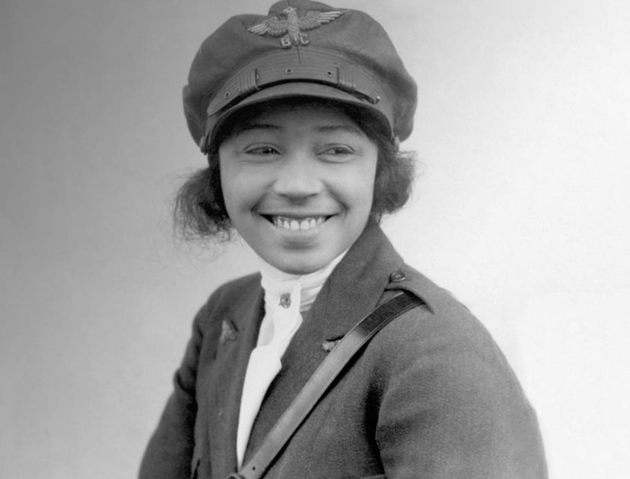 Bessie Coleman, a woman who appears to be in her late 20s, smiles at something to the right of the camera. She is wearing a military-like uniform, with an emblem that reads "B.C." on her cap.