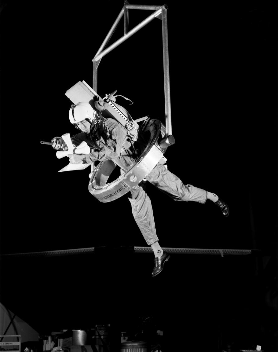 In this black and white image, a man is suspended from an apparatus while using his hands to handle a device. 