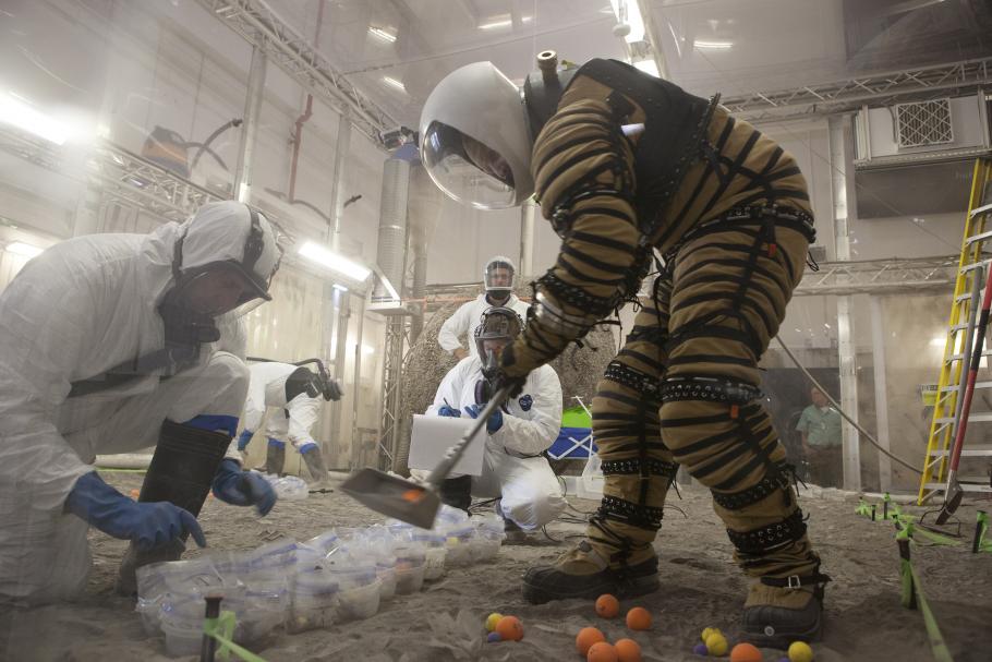 A person outfitted in the new spacesuit uses a tool to pick up balls and place them in bags in front of him. 