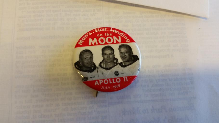 Pin with image of Apollo 11 astronauts in the center and at top and bottom white text against red background. 