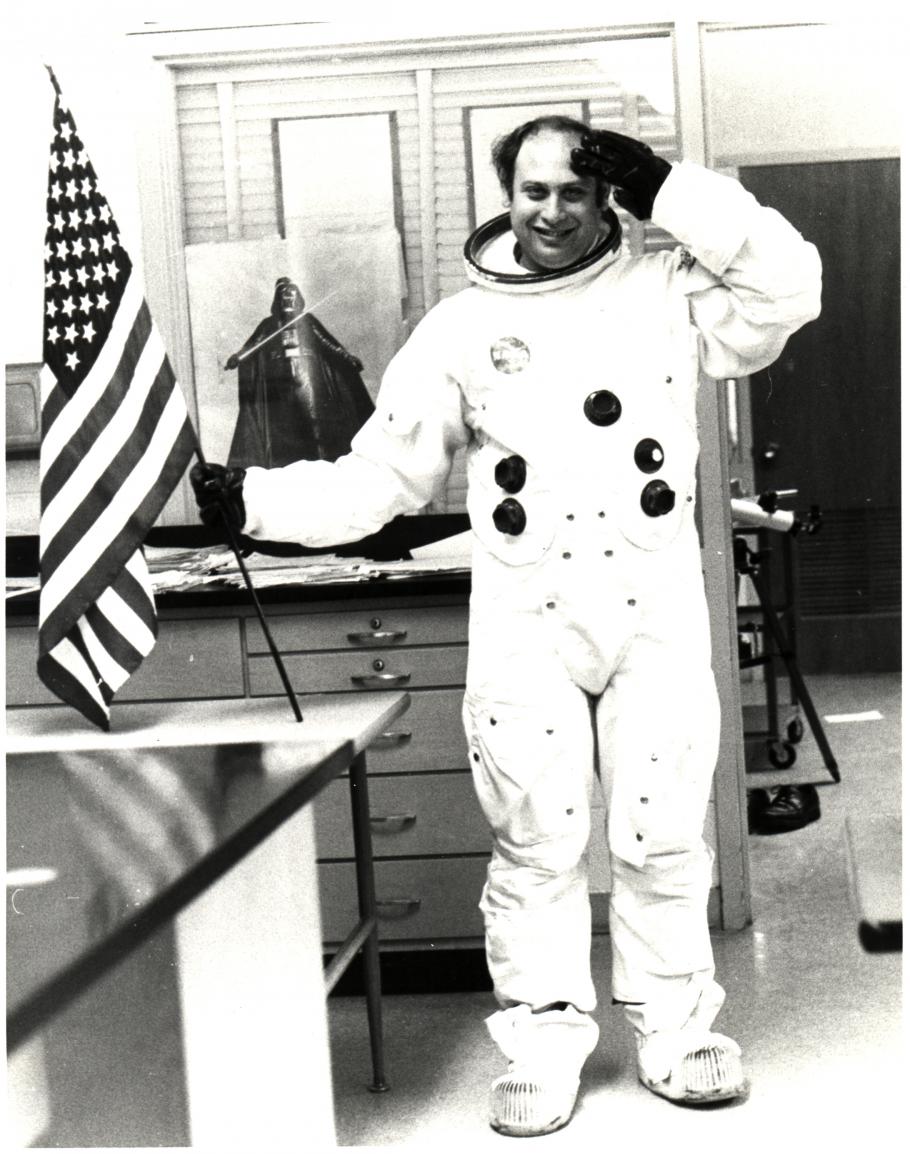 Man wearing space suit without a helmet salutes with his left hand while holding American flag in his right