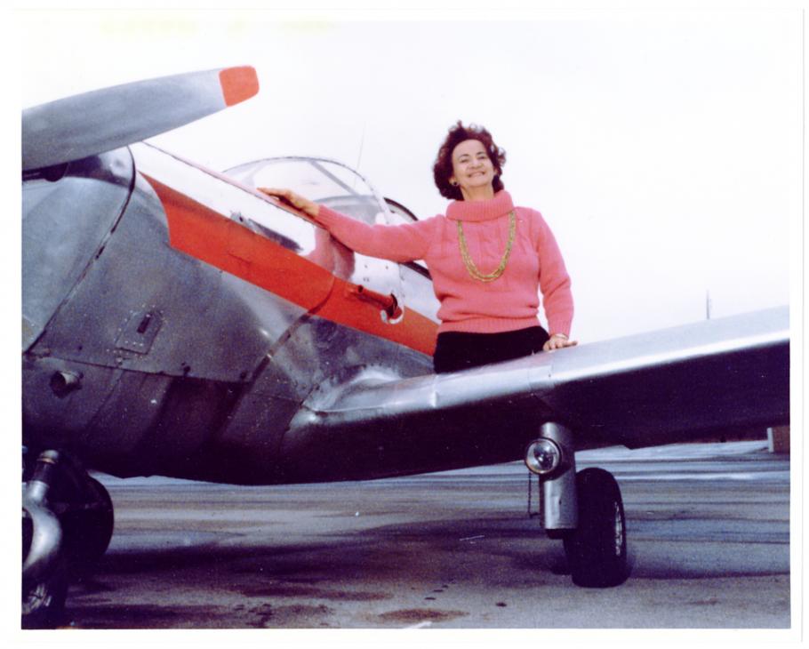 A partial view of the left side of an airplane with a white then red stripe under the cockpit. A woman in a pink mock turtleneck sweater with a gold chain necklace sits on the wing of the airplane. She has lost both legs above the knee.