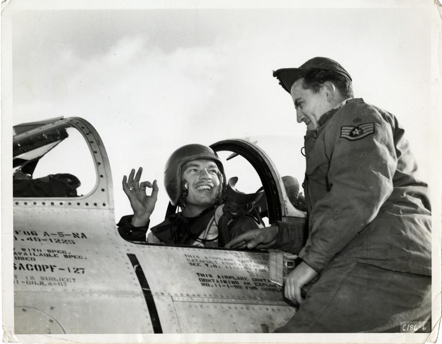 Man in a helmet sits in an aircraft cockpit giving the okay sign with his right hand to a man in a cap and flight suit on his left