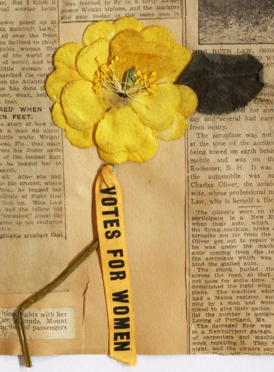 Artificial flower, bright yellow double layer petals attached to light green wire stem; below stem yellow ribbon with black printed text "VOTES FOR WOMEN"