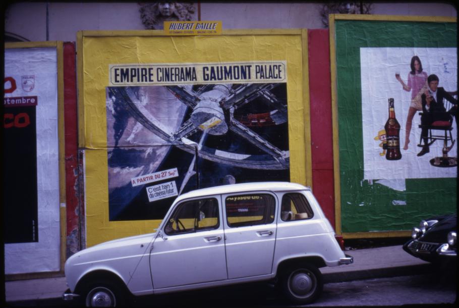 A white vehicle is parked in front of a movie poster for the film "2001: A Space Odyssey" plastered on multicolored wall. 