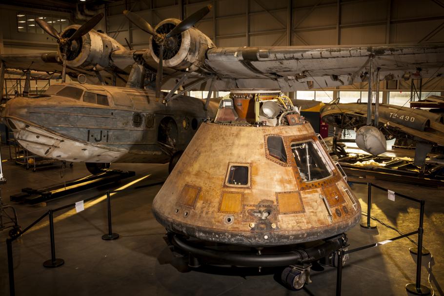 A conical shaped structure, which is the Apollo Command module, sits in front of an aircraft with signs of age.
