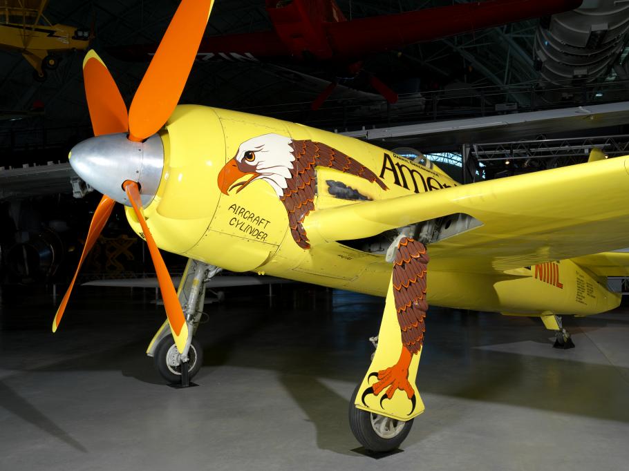 Side view of front of yellow monoplane with orange, four-blade propellor attached to a single engine. Artwork of an eagle is painted throughout the visible side of the aircraft. Aircraft is on display at the museum.