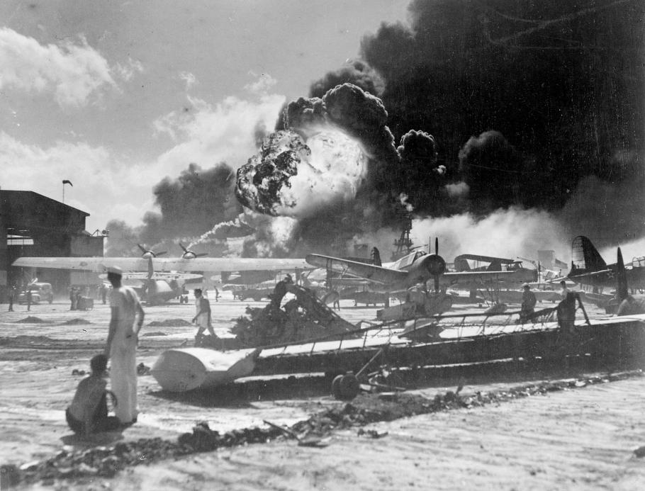 Sailors stand in the foreground. Damaged aircraft and explosions appear in background. 
