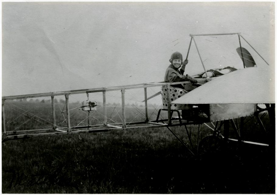 Rearview shot of an early airplane with Moisant in cockpit looking over her shoulder and smiling at the camera. 