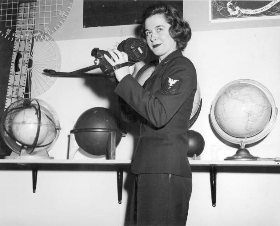 Woman in uniform stands in front of a series of globes and holds a link sextant.