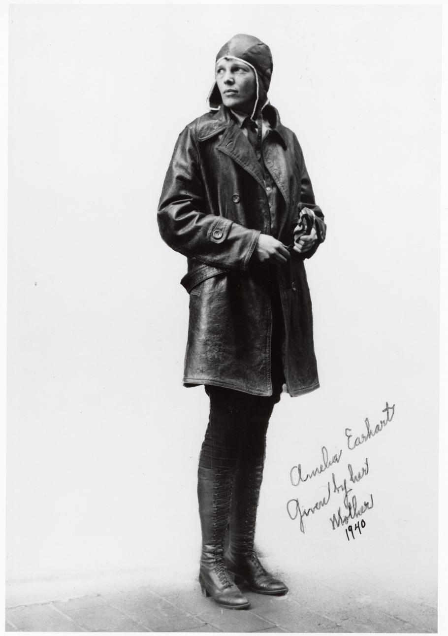 Amelia Earhart, a woman, poses for a photo in aviator gear. Her name is written in cursive script next to her legs.