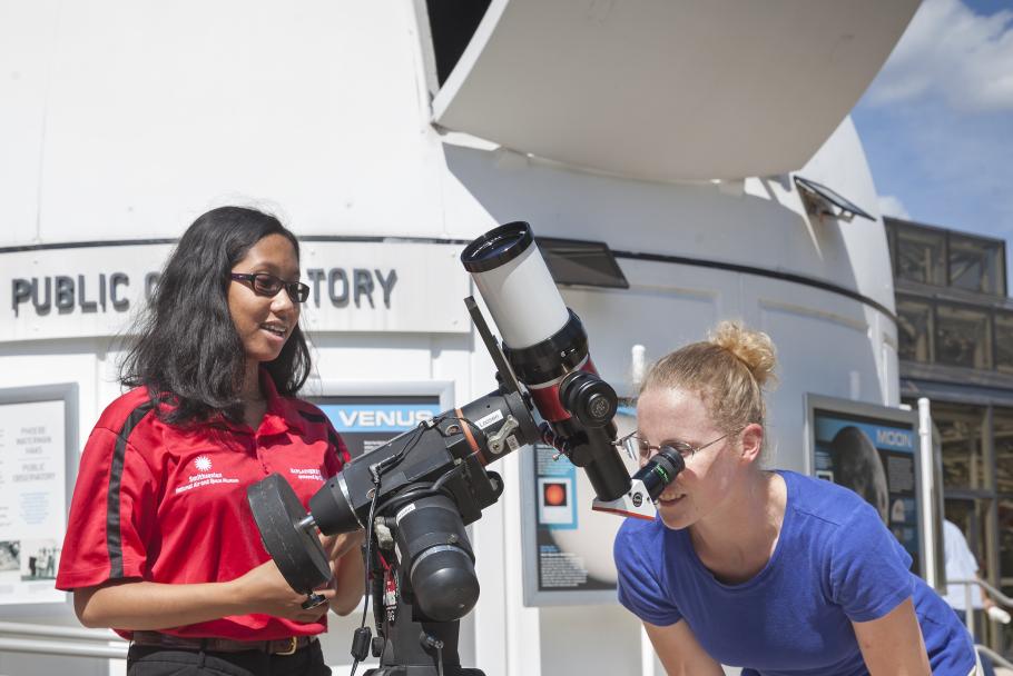 A woman looks in a telescope while a person in a polo stands by. 