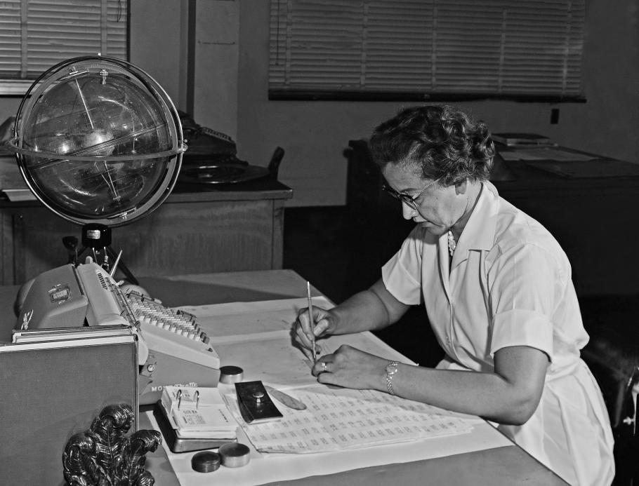 A woman in a collared shirt and necklace leans over her desk working.