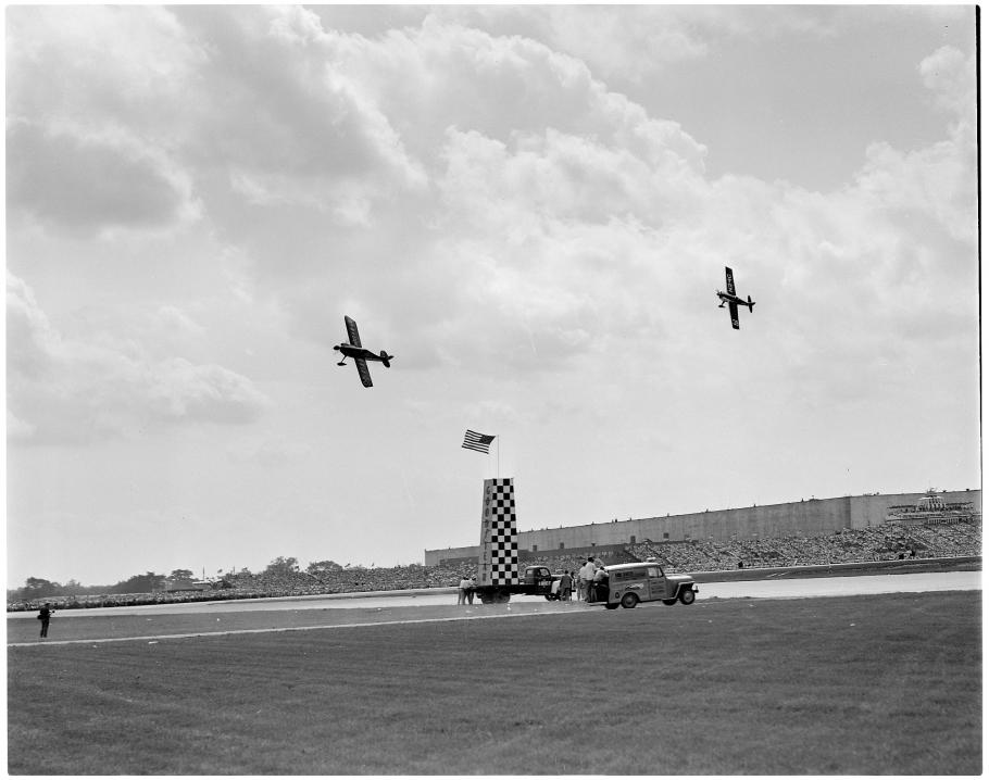 Two flying aircraft rounding a pylon adorned with the American flag at a race track. People sit in the stands in the far background.