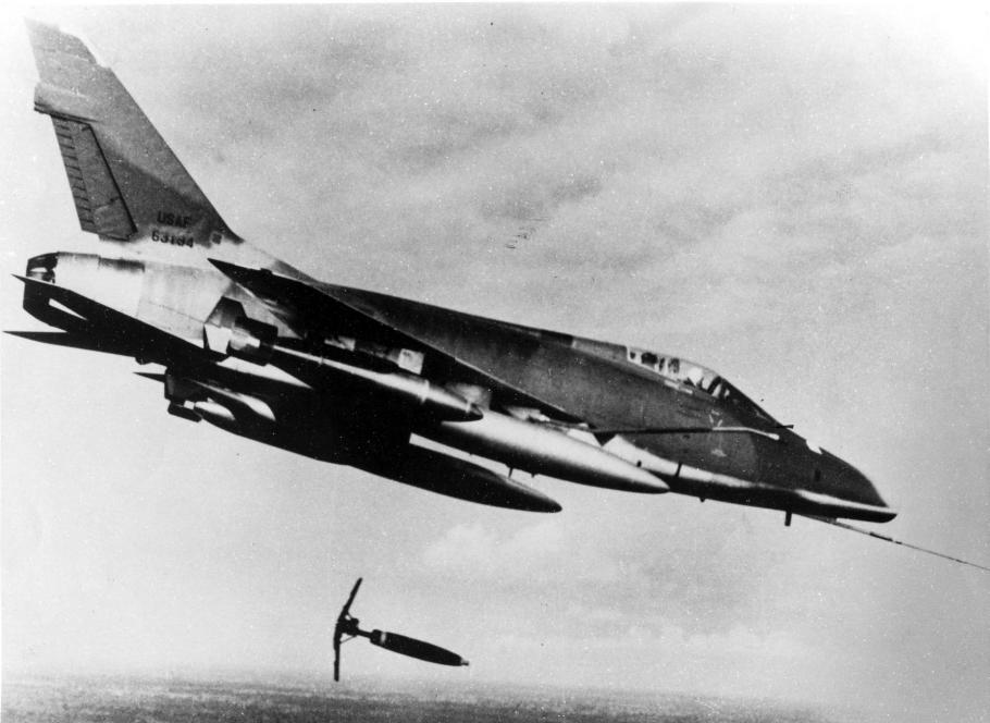 A black and white photo of a North American F-100D aircraft in flight dropping a "Snake eye" bomb on a suspected Viet Cong position in 1966. 
