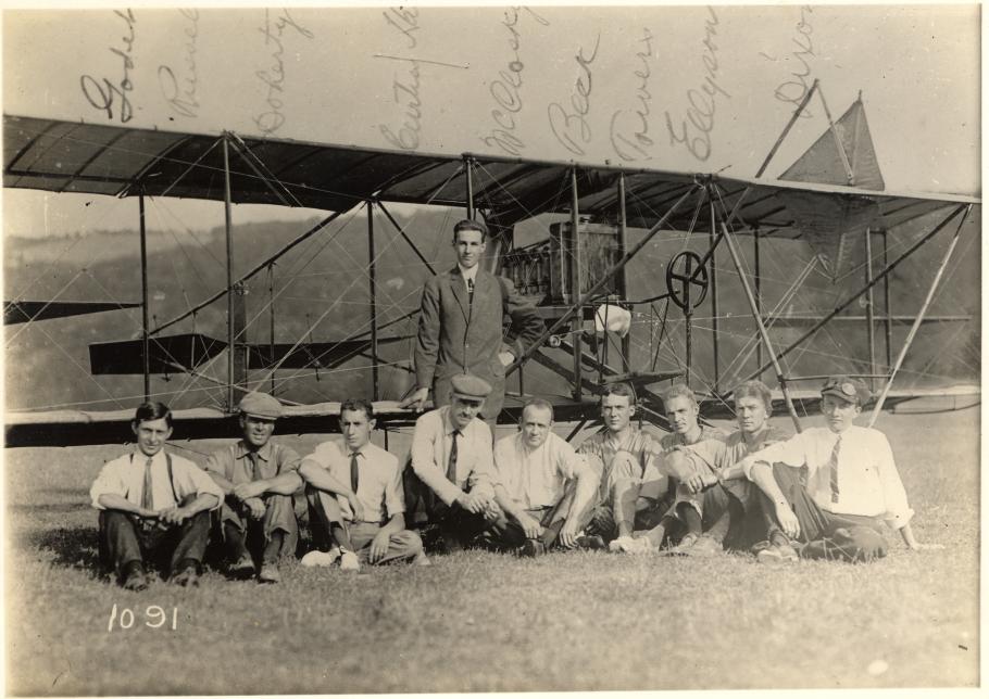 Eight men sitting, one man standing in front of airplane