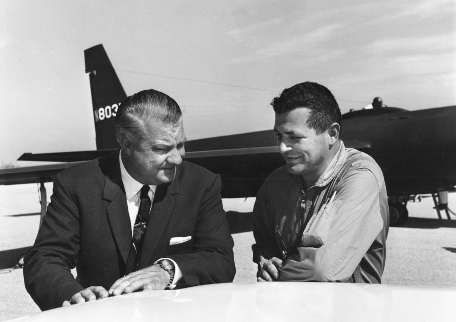 Two men, one in a suit, one in a flight suit, stand in front of an airplane.