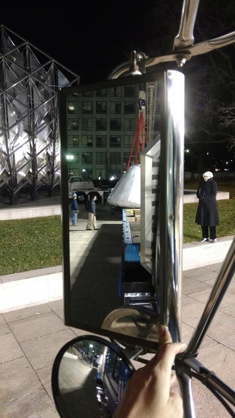 Image of a rear view mirror. In the mirror you can see the outline of the command module on a truck flat bed. 