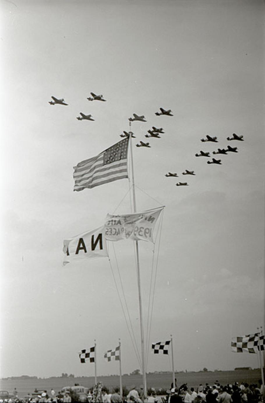Formation of airplanes fly past American flag. Spectators can seen at bottom.