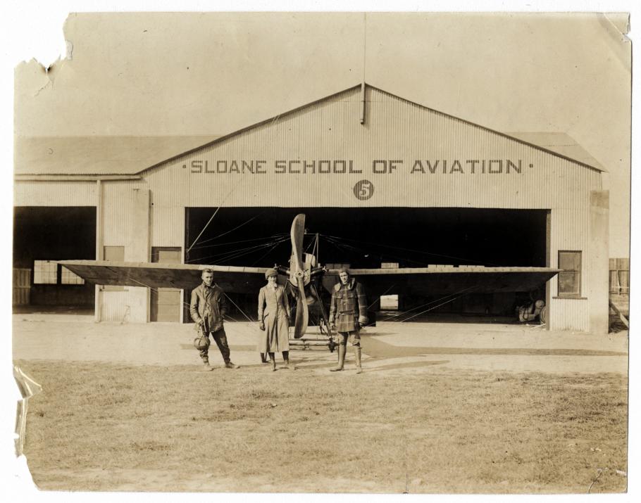 Two men and a woman in front of a hangar and airplane