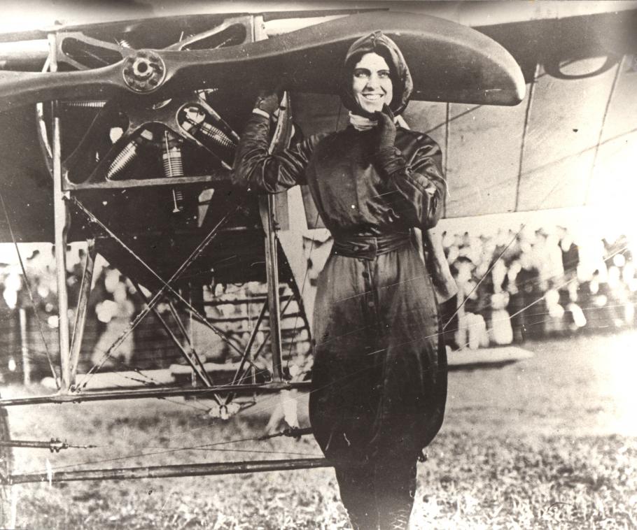 Woman in flying gear poses in front of an airplane. Right hand on propeller, left hand on her chin. Crowd in background.