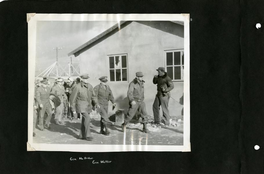 Black scrapbook page. Centered photo in photo corners: 3 men in uniform (Gen. Douglas MacArthur 3rd on the left) walk in front of a building. Background left: group in uniform following. Background right: Man in leather jacket and cap.