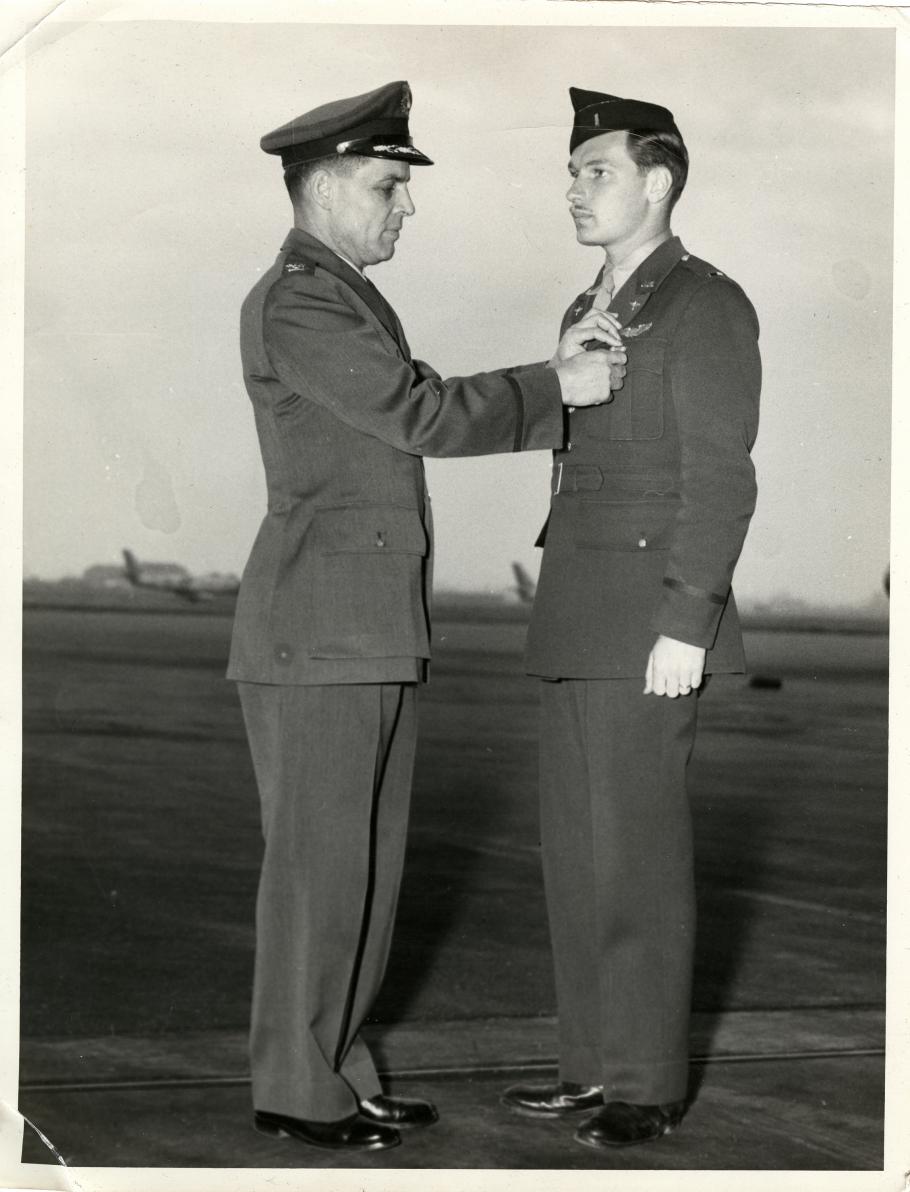 (Left) Man in military uniform and hat pins an award on man in cap on right. Air field with aircraft in background.