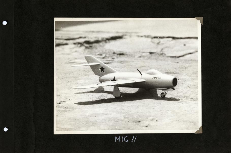 Black scrapbook page with photo of the right side of model airplane with star on tail and fuselage centered. Handwritten white text below reads: "MIG!"