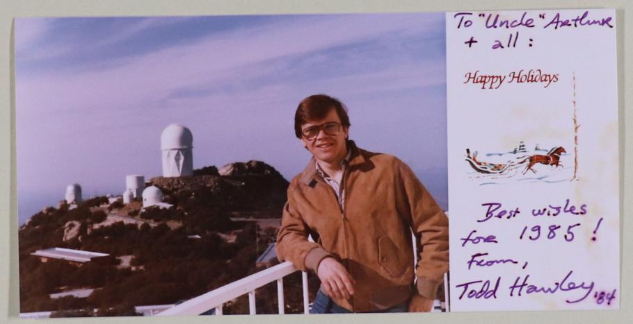 (left) Man with brown hair and glasses, wearing a tan jacket, stands at a railing. Background is an observatory with white buildings on a hill. (right) preprinted message "Happy Holidays" with drawing of horses and sleight. Handwritten note.