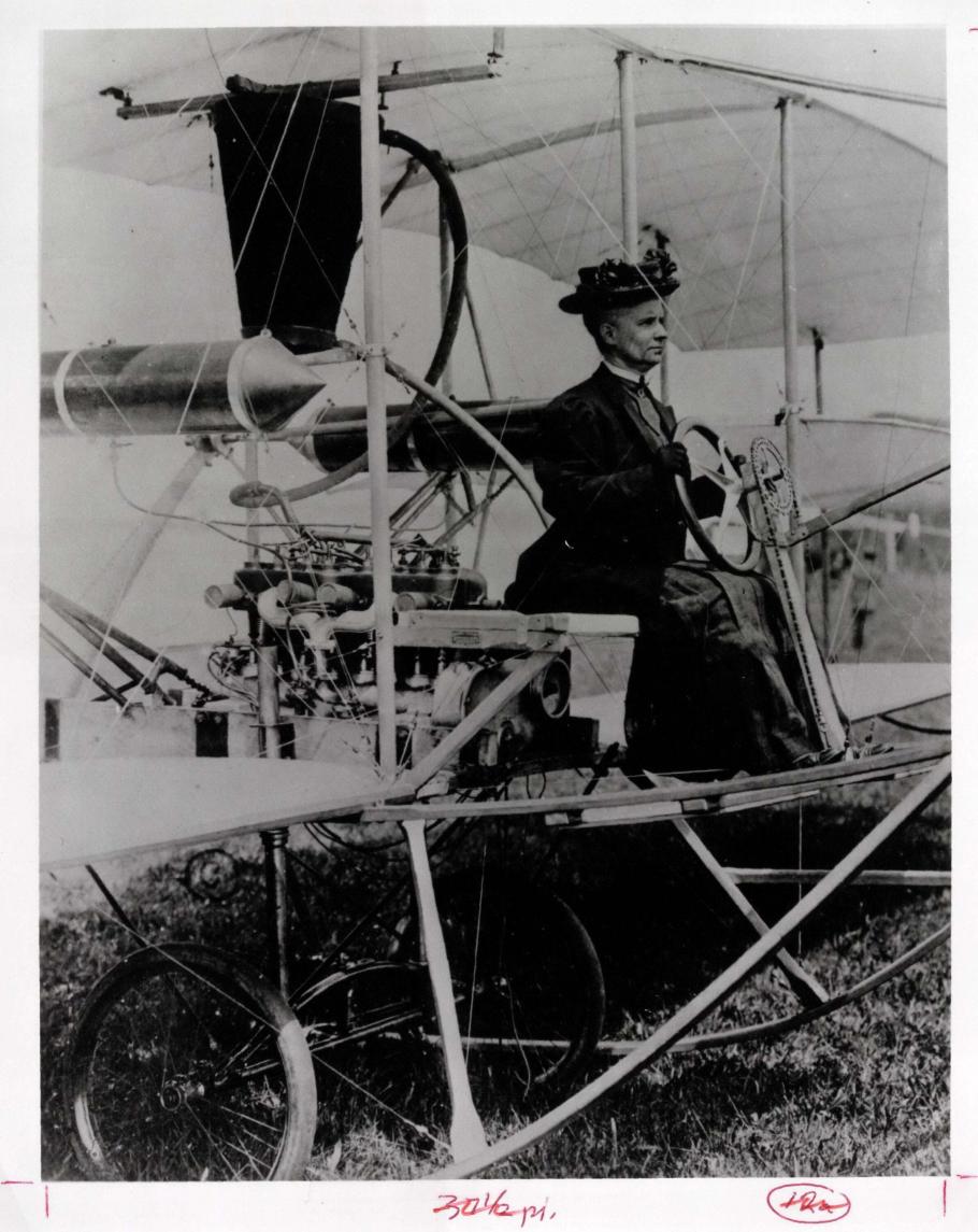 Woman seating at the steering wheel of an early airplane, wearing a hat