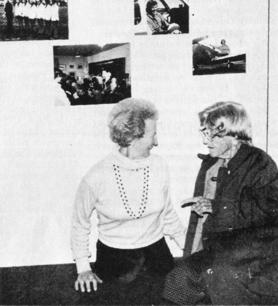 Black and white photograph of two women in front of a gallery wall with four photographs. Woman on the left wears a white shirt and a necklace. Woman on the right wears a dark jacket.