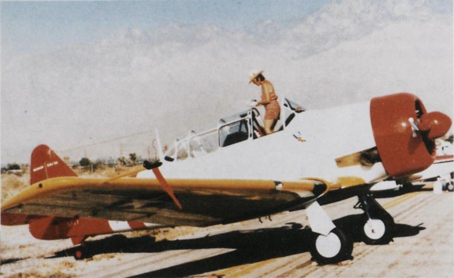Right side view of woman in hat and khaki shorts stands in the cockpit of a white aircraft with yellow wings and red nose and tail. Front wheels are down.