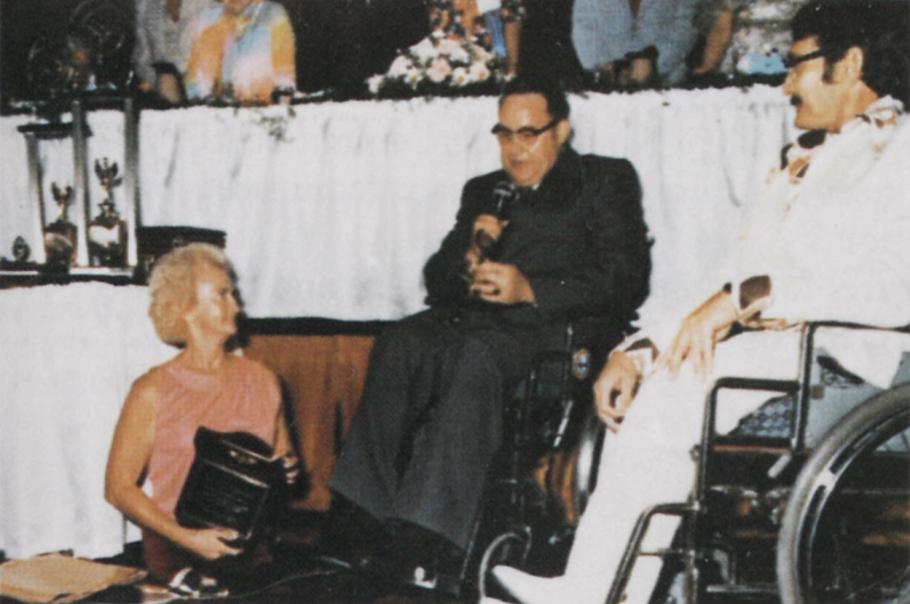 From right to left: woman in pink shirt holds a plaque (she is missing both legs and walking on her knees). Man wearing glasses and a black suit holds a microfilm, sitting in a wheelchair. Man in white suit sitting in wheelchair. Backdrop is banquet table