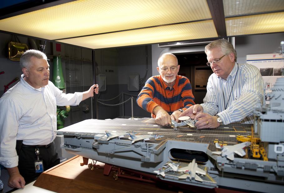 curator Chris Moore (left) looks on as Steve Henninger (center), assisted by Paul Moore, frees the E-2C on the model of the USS Enterprise.