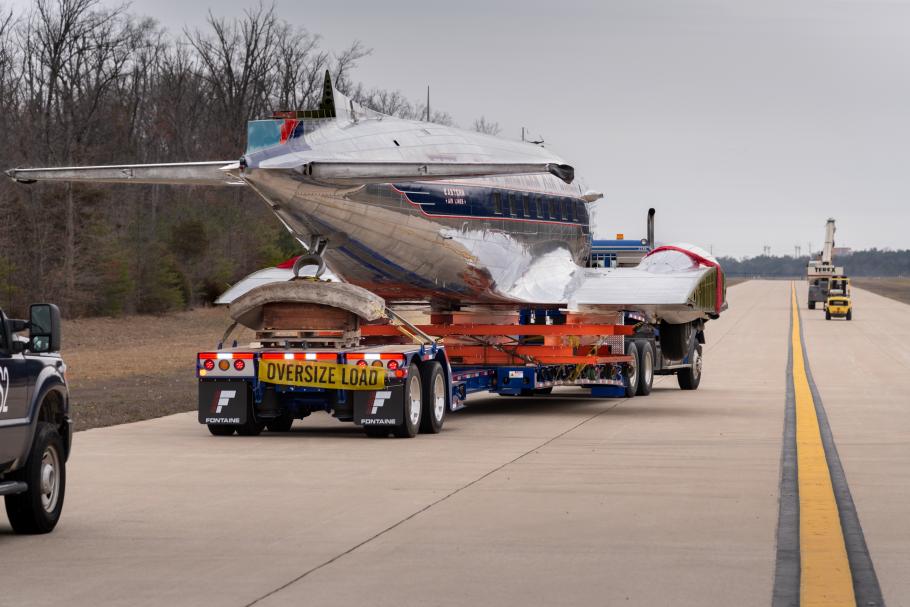 The Douglas DC-3 is on a truck on a highway. It is moving away from the camera. 