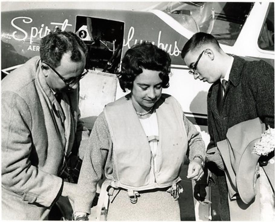 Black and White photograph: Airplane in the back has writing: Spirit of Columbus. Woman in the center has dark hair and wears and inflatable vest. Men on left and right wear glasses and are turning towards the woman.