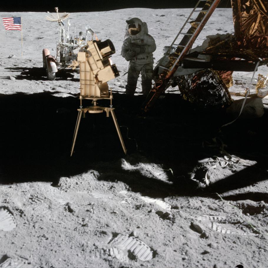 Image of camera on the Moon; Lunar Module, Astronaut, and American flag in background. 